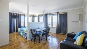 Una sala moderna con piso de madera, un sofá de cuero negro, una mesa de comedor con sillas y una cama con colcha azul.