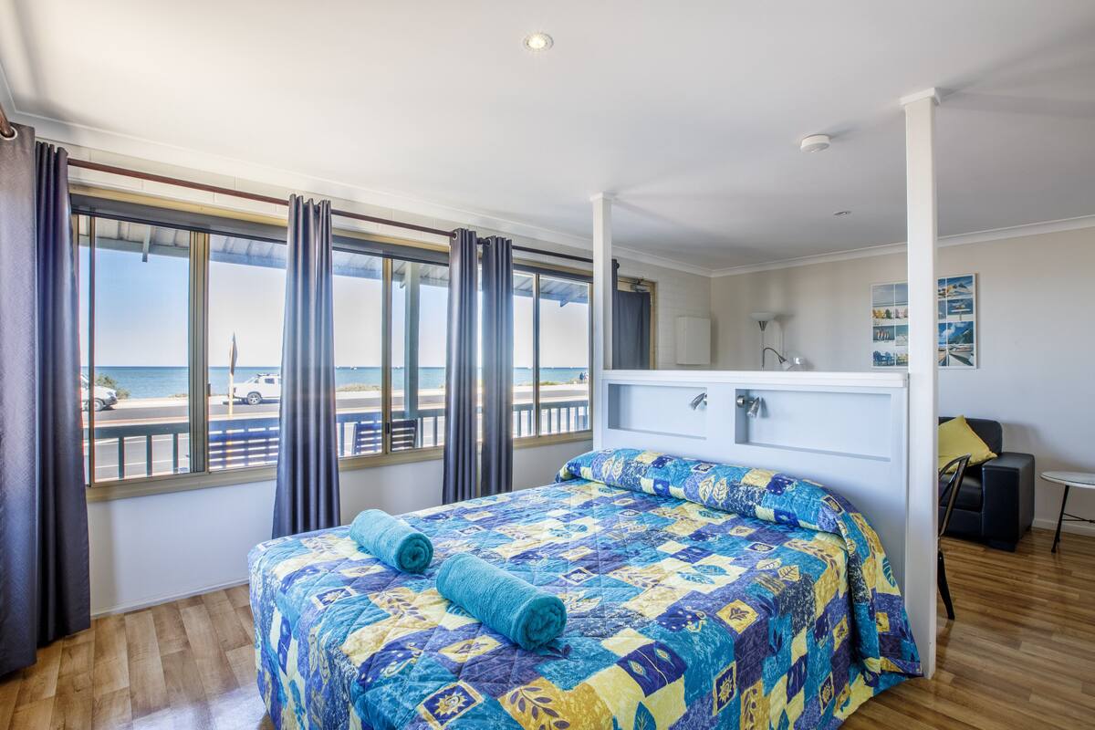 Un dormitorio con una cama grande, vista a la playa y un escritorio pequeño con una silla.