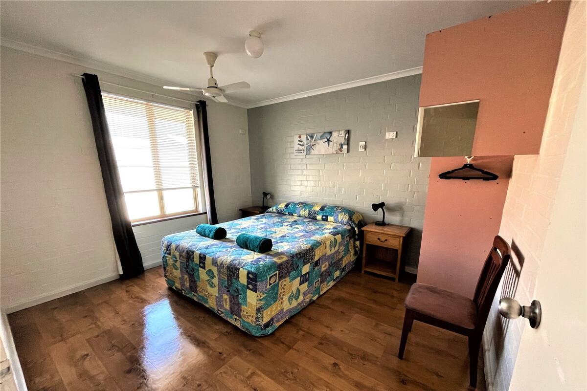 Un dormitorio con piso de madera, una cama con colcha colorida, un ventilador de techo, una mesita de noche y una silla.