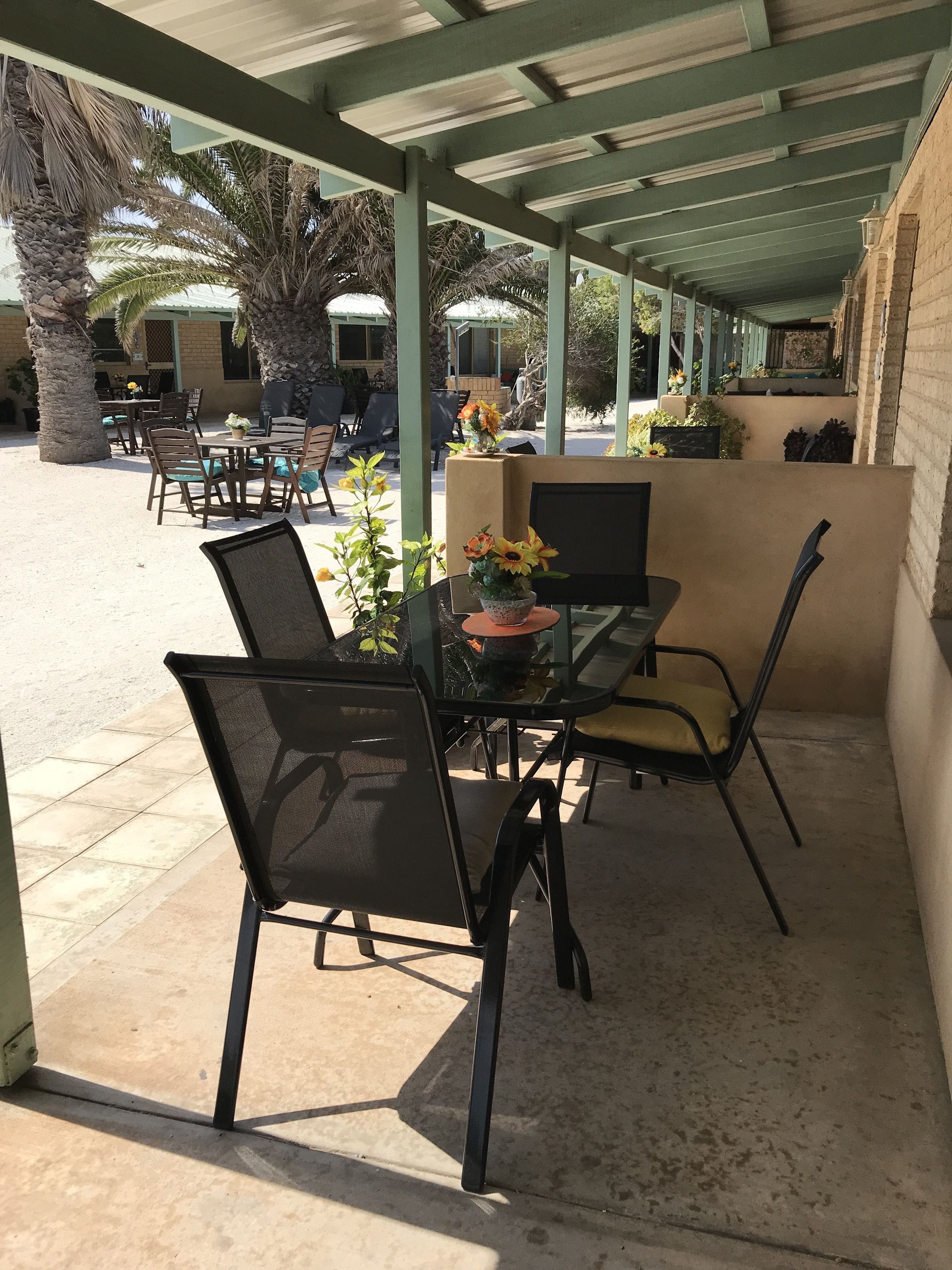 Un patio al aire libre con una zona de estar cubierta, que incluye una mesa y sillas, rodeado de palmeras y plantas en macetas.