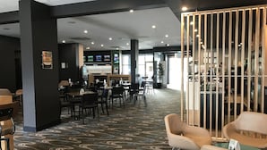 Un moderno vestíbulo de hotel con alfombra estampada, zonas de estar, un bar y una pantalla grande mostrando contenido.