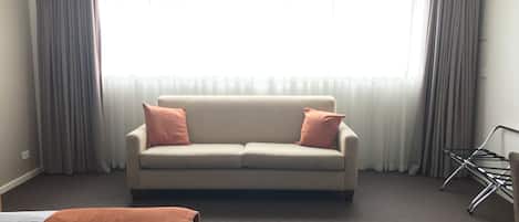 A hotel room with a beige sofa, two pillows, a bed with a brown cover, a chair, and a window with sheer curtains.