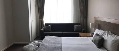 A hotel room with a large bed, a sofa, a nightstand, and a window with curtains.