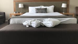 Una cama bien tendida con sábanas blancas, dos lámparas de noche, un cabecero gris y un par de toallas dobladas sobre un otomano oscuro.