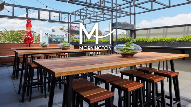 Un área de estar exterior moderna con mesas y taburetes de madera, un recinto de vidrio y un letrero del Mornington Hotel.