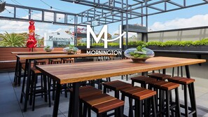 Un área de estar exterior moderna con mesas y taburetes de madera, un recinto de vidrio y un letrero del Mornington Hotel.