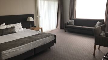 A hotel room with a large bed, a sofa, a chair, a nightstand, and a lamp.