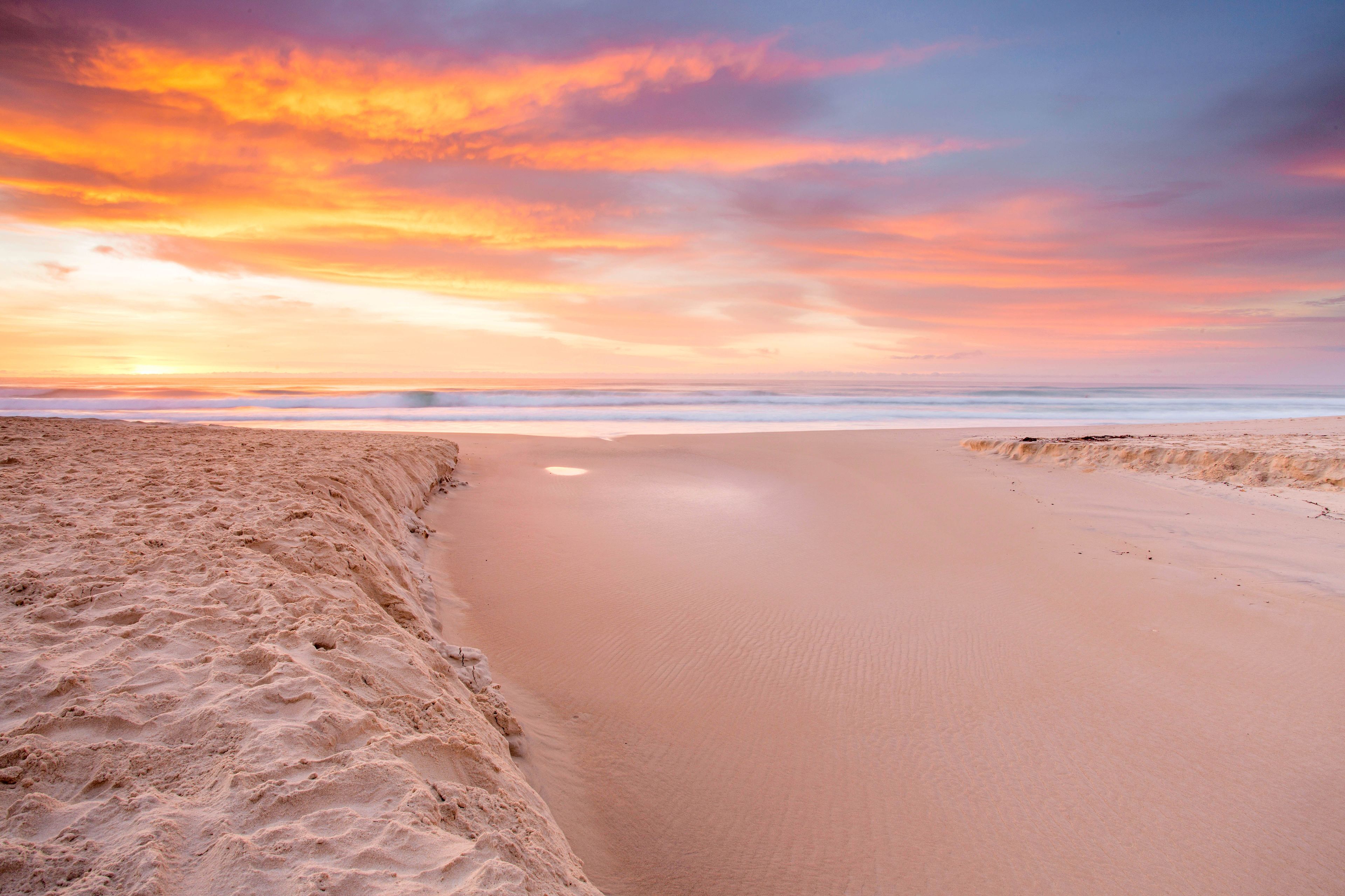 A beach with a wide sandy shore and a calm sea under a colorful sunset sky.