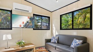 Una sala de estar moderna con un sofá gris, una mesa de centro de madera y una alfombra colorida. Hay ventanas grandes con vistas tropicales y un equipo de aire acondicionado montado en la pared.