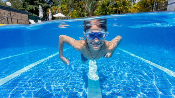 Vista subacuática de una piscina con agua azul cristalina, suelo de baldosas y un nadador.