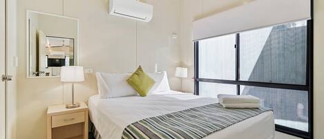 A hotel room with a bed, bedside lamps, a nightstand, a mirror, an air conditioning unit, and a large window.