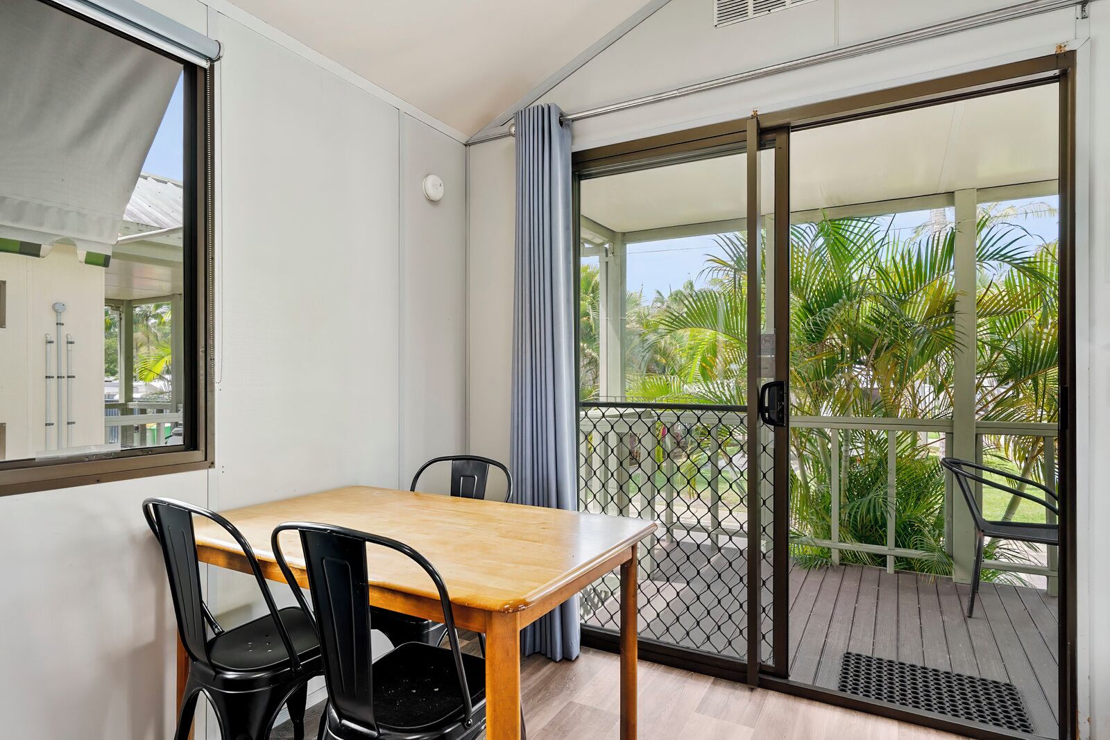 A room with a wooden table and chairs, a sliding glass door leading to a balcony with a view of palm trees.