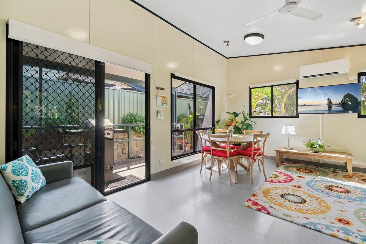 A modern living room with a sofa, dining table, chairs, and a bench. There is a sliding glass door leading to an outdoor area with a barbecue grill.