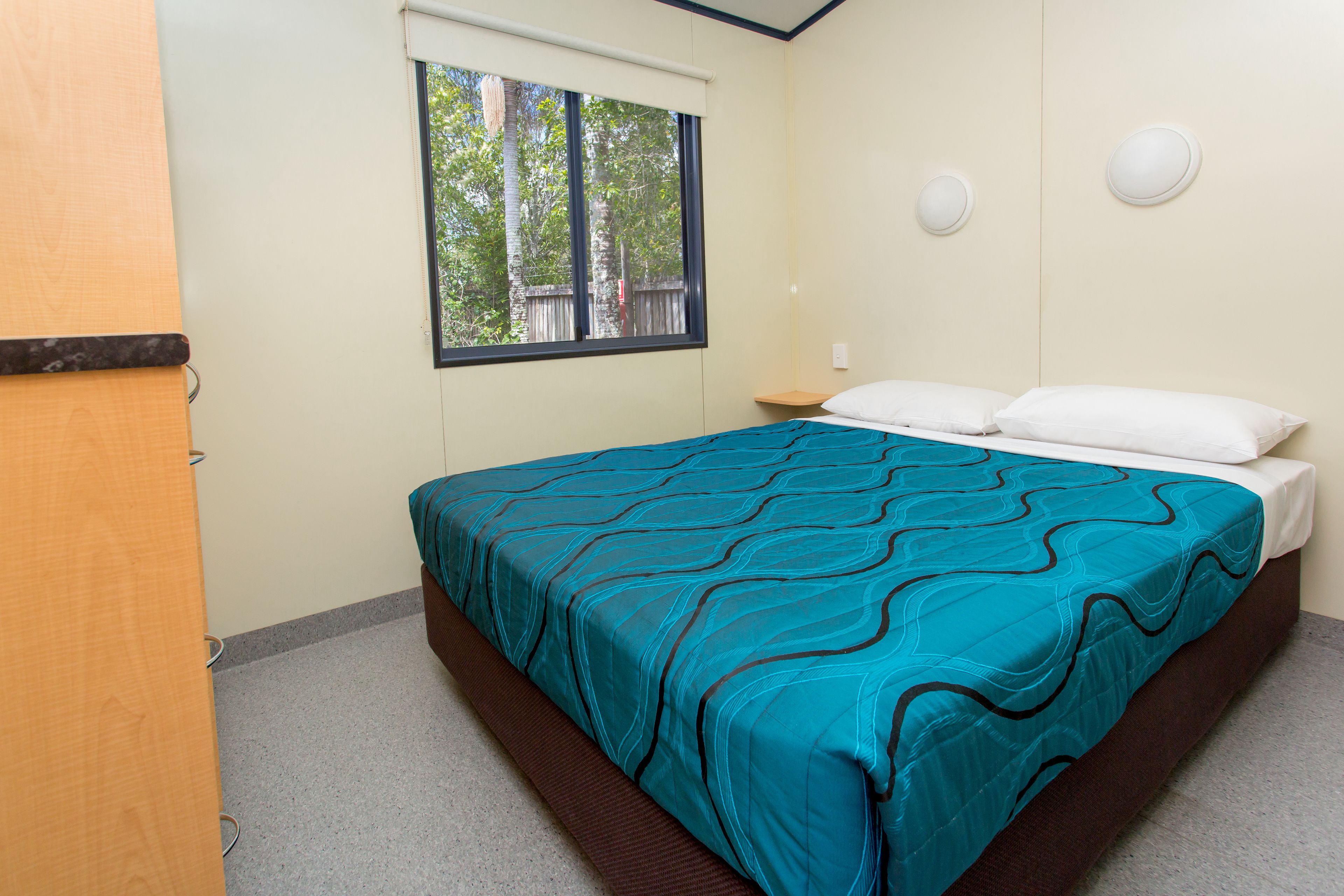 A bedroom with a bed, a window, a nightstand, and a wall-mounted light fixture.