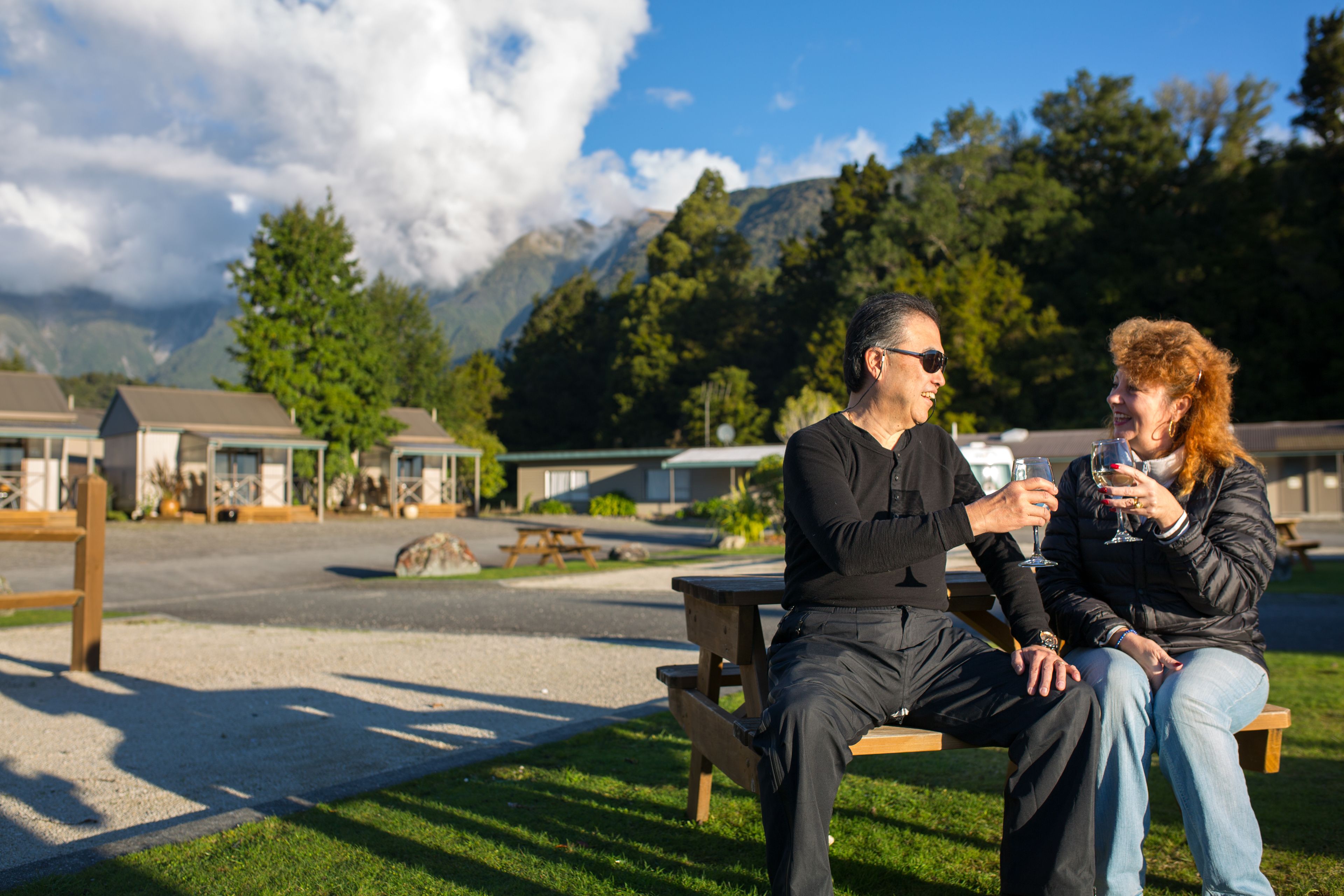 Two people sitting on a wooden bench outdoors with a mountainous backdrop.