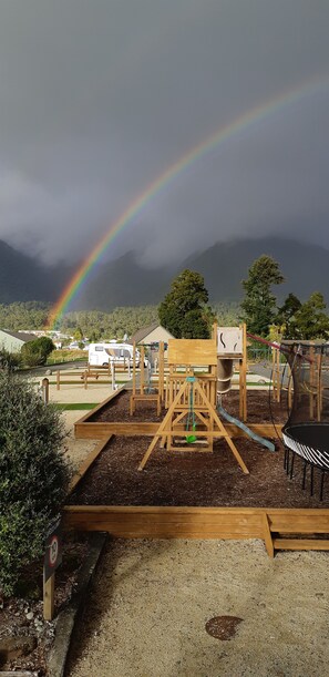 A playground with wooden structures and a dirt ground, surrounded by trees and a mountain in the background. - Fox Glacier TOP 10 Holiday Park & Motels (Fox Glacier)
