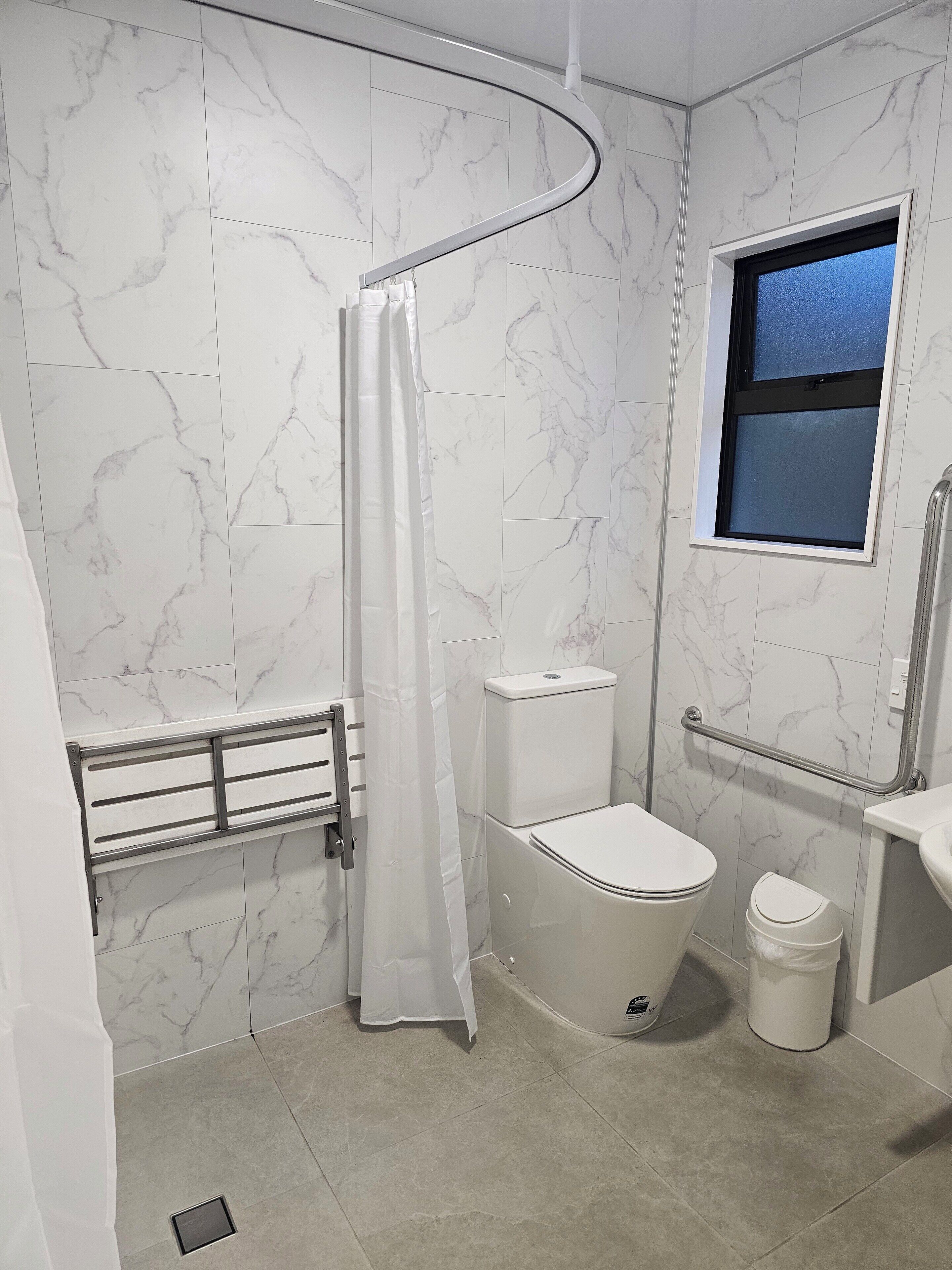 A bathroom with a white toilet, a shower with a white curtain, a grab bar, a small window, and a white trash bin.