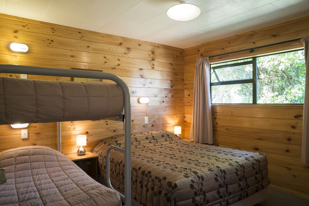 Habitació amb parets de fusta, un llit superposat, una taula de nit amb una làmpada i una finestra amb cortines.