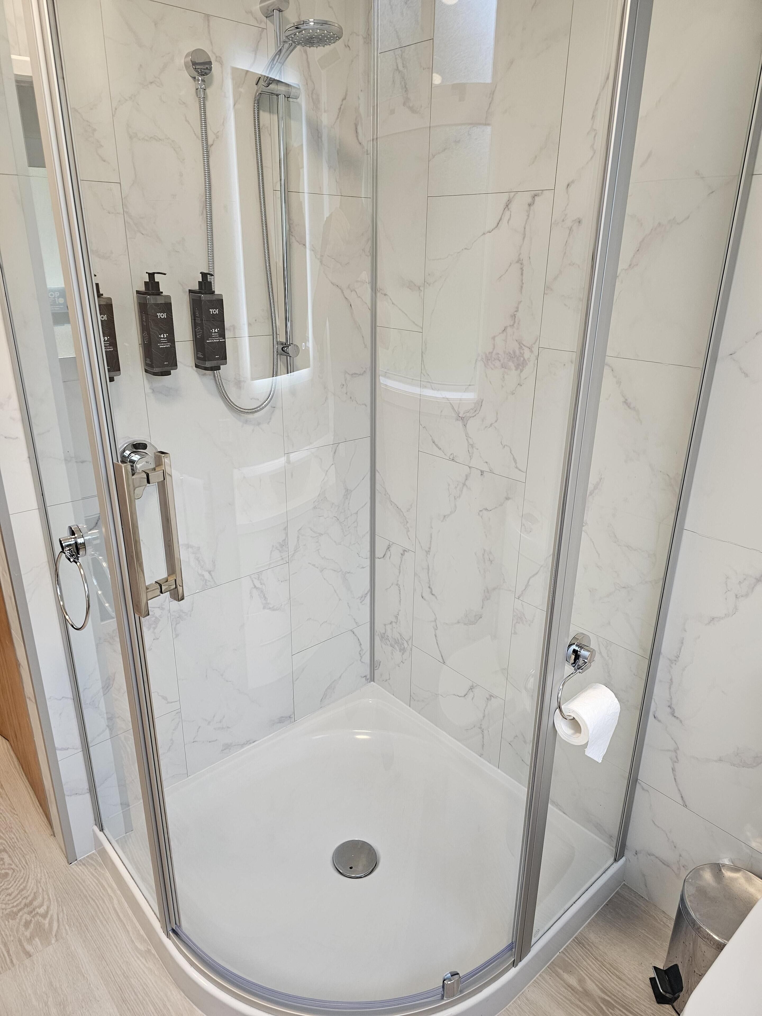 Une douche fermée par des parois de verre, avec un mur recouvert de carreaux de marbre, une douchette à main et un distributeur de savon à main.