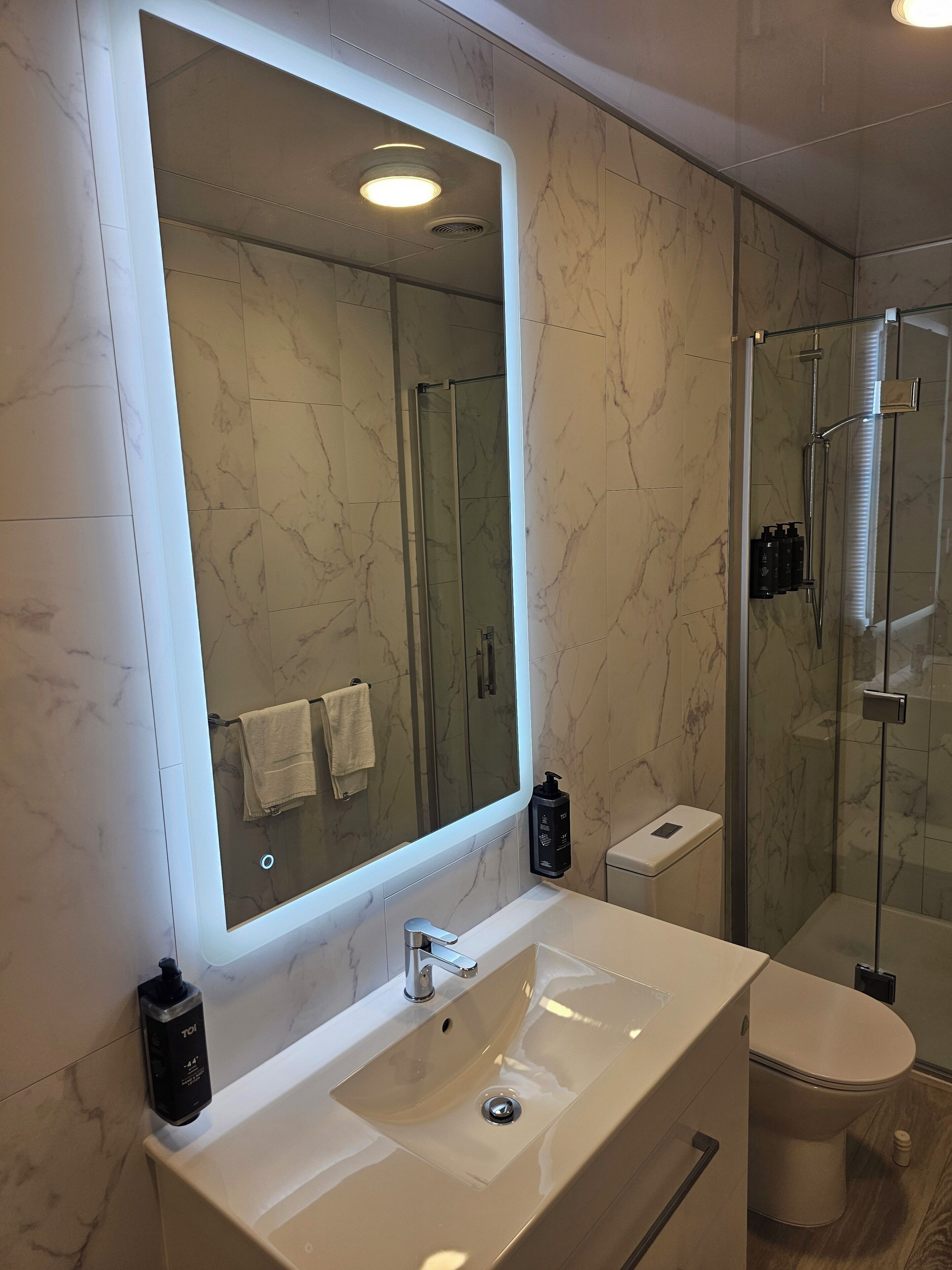 A modern bathroom with a large mirror, white marble tiles, a glass-enclosed shower, and a white sink with a soap dispenser.