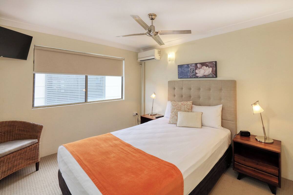 A hotel room with a bed, a chair, a ceiling fan, a window with blinds, an air conditioning unit, and a painting on the wall.