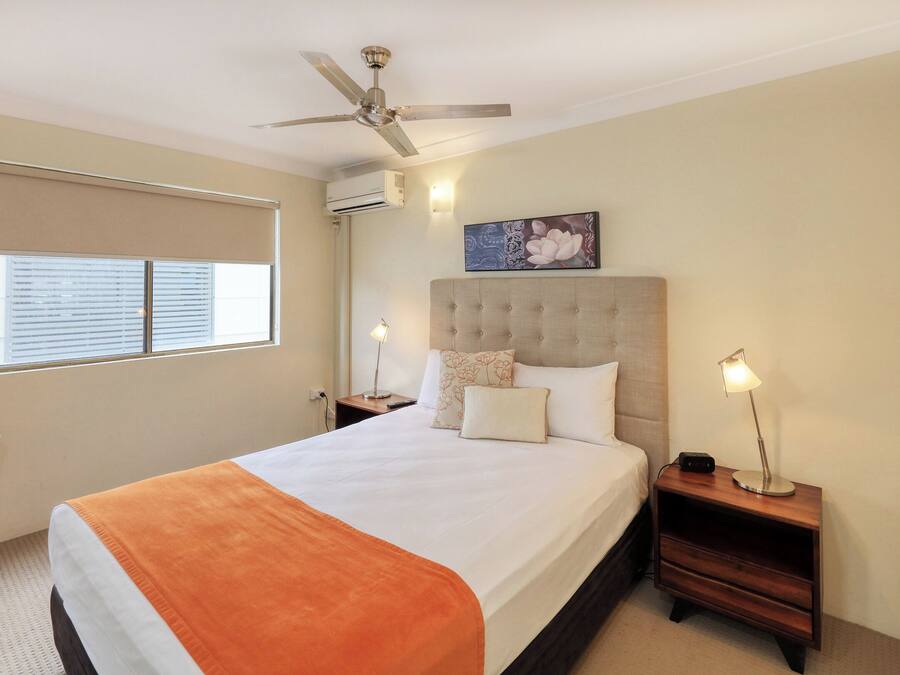 A hotel room with a bed, a chair, a ceiling fan, a window with blinds, an air conditioning unit, and a painting on the wall.