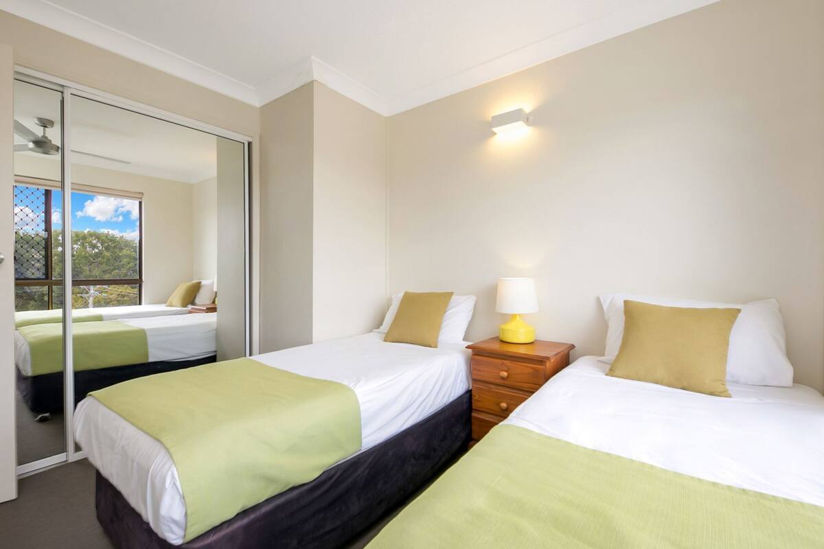A hotel room with two beds, a mirror, a nightstand with a lamp, and a view of trees through the window.