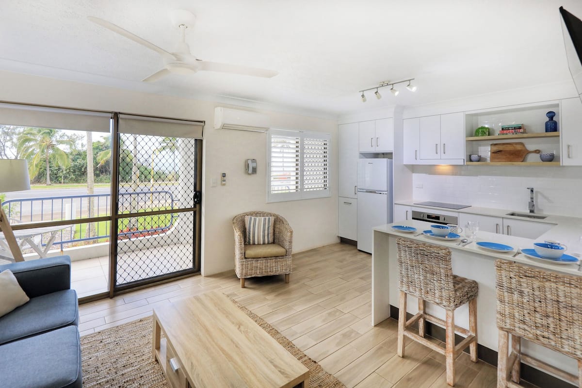 A modern kitchen with white cabinets, a dining area with wicker chairs, and a balcony with a view of palm trees.