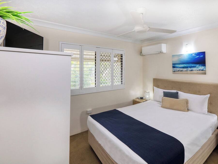 Un dormitorio con cama, mesitas de noche, ventilador de techo, aire acondicionado, ventana con persianas y televisor de pared.