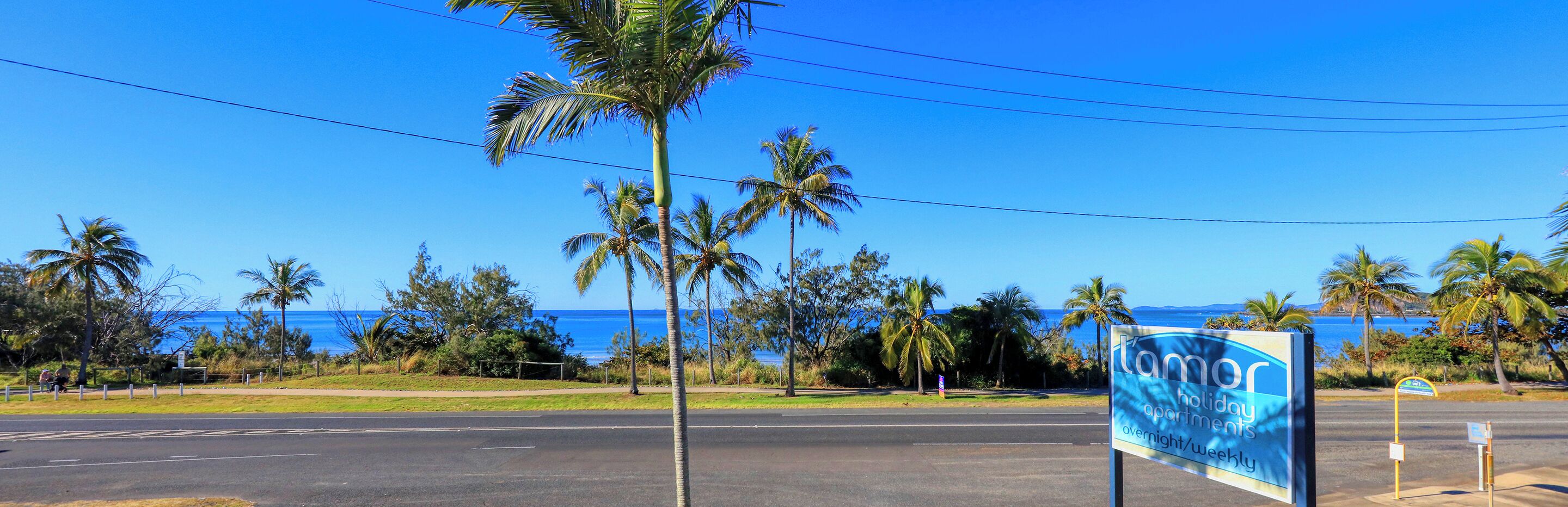 A sign for 'Lamor Holiday Apartments' with a view of the ocean and palm trees.