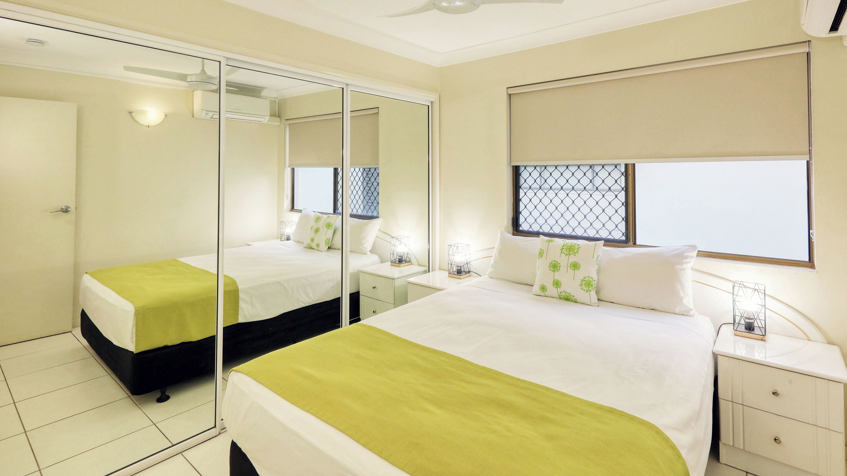 Une chambre d’hôtel avec un grand lit, un lit plus petit, une armoire, une table de chevet, une commode, une fenêtre avec un store et un ventilateur de plafond.