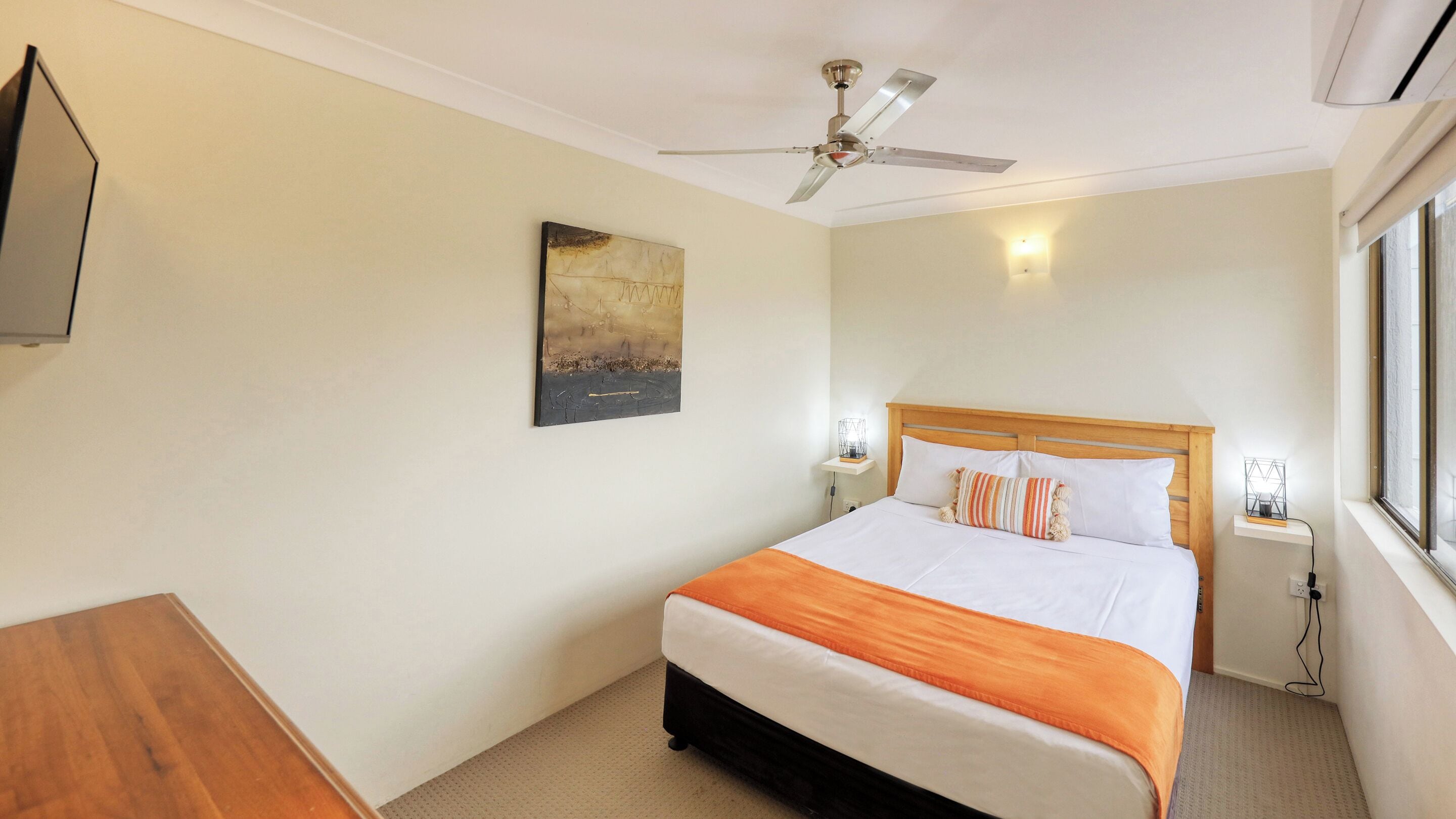 Une chambre d’hôtel avec un lit, un ventilateur de plafond, un tableau au mur, une table de chevet avec une lampe et une fenêtre.