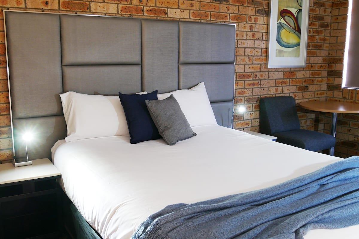 A hotel room with a brick wall, a large bed with white bedding, two pillows, a grey headboard, a small round table, and a blue chair.