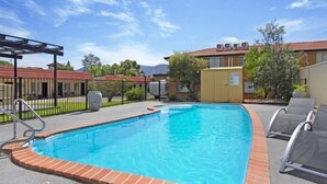A rectangular swimming pool with a brick edge, surrounded by a paved deck. There are two lounge chairs on the right side. - Country Capital Motel (South Tamworth)