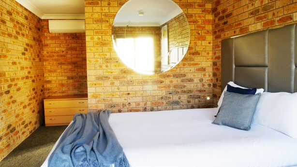 A bedroom with a brick wall, a round mirror, a bed with pillows, and a nightstand. - Country Capital Motel (South Tamworth)