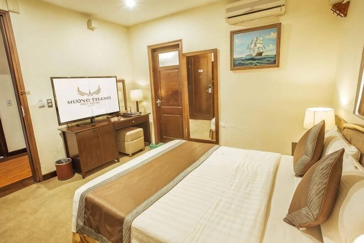 Quarto de hotel com cama, escrivaninha, televisão, espelho e um quadro na parede.