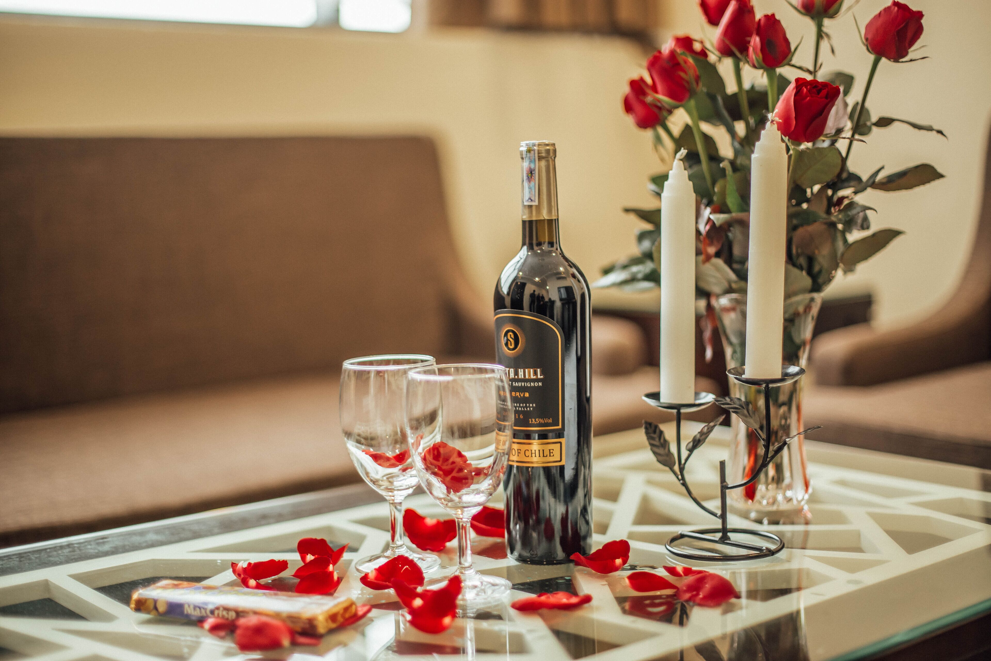 Una mesa de vidrio con una botella de vino, dos copas y pétalos de rosa.