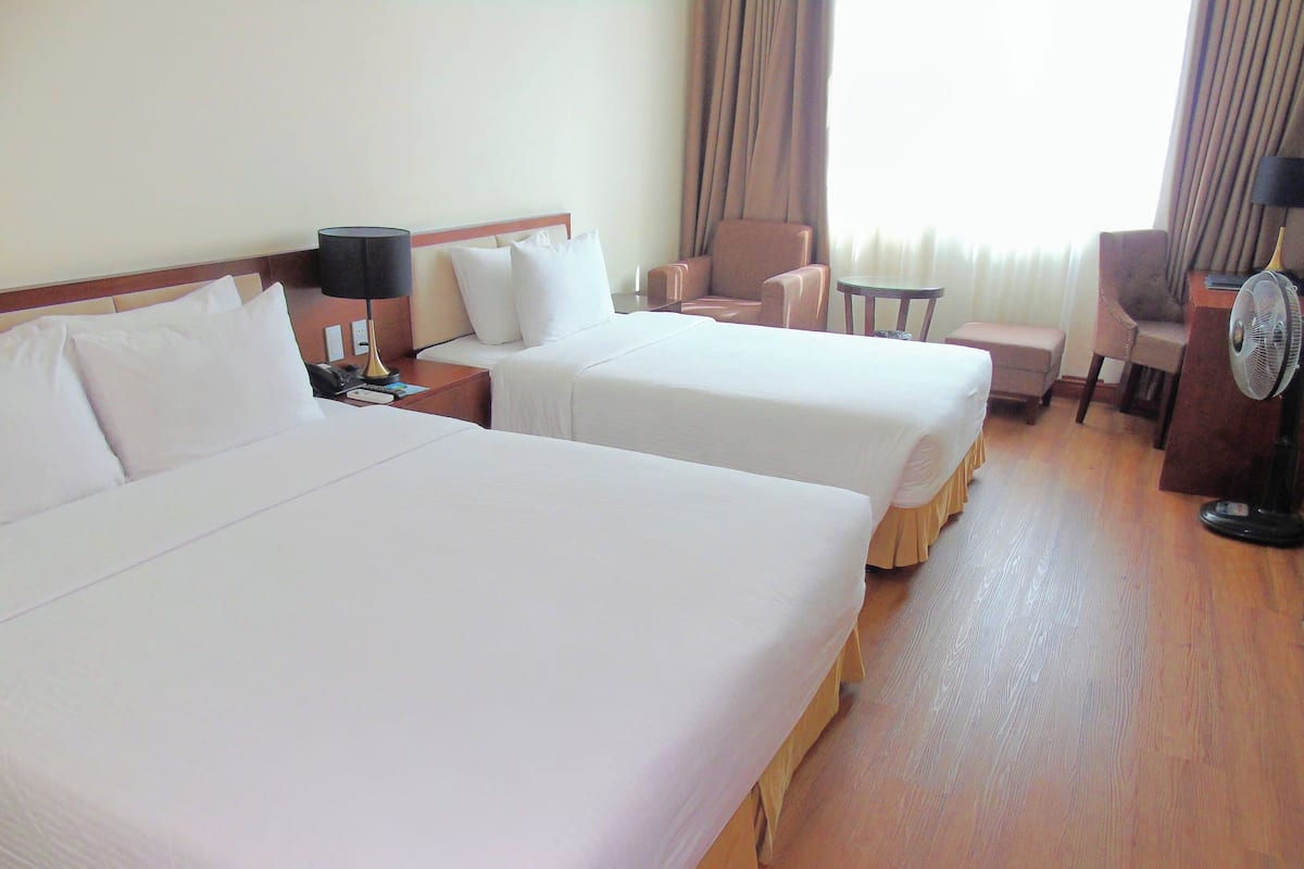 Une chambre d’hôtel avec deux lits simples, une table de chevet avec une lampe, une chaise, une petite table et un ventilateur sur pied.