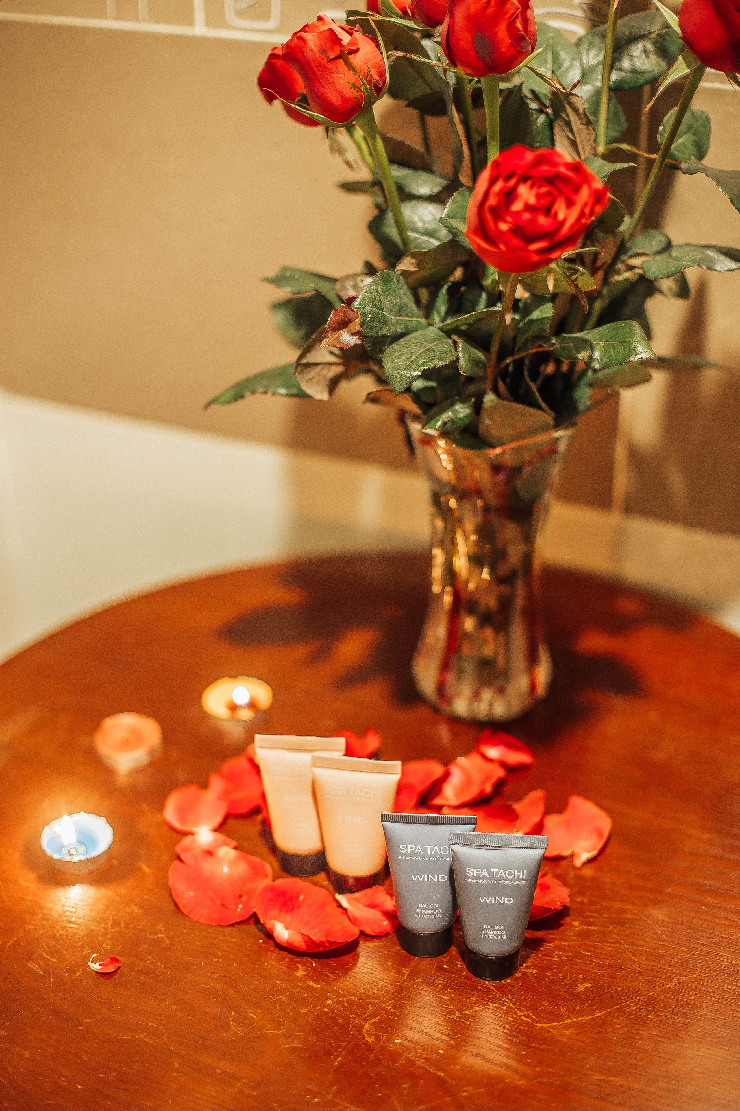 Una mesa con un jarrón de rosas rojas, dos tubos de productos de spa y pétalos de rosa esparcidos.