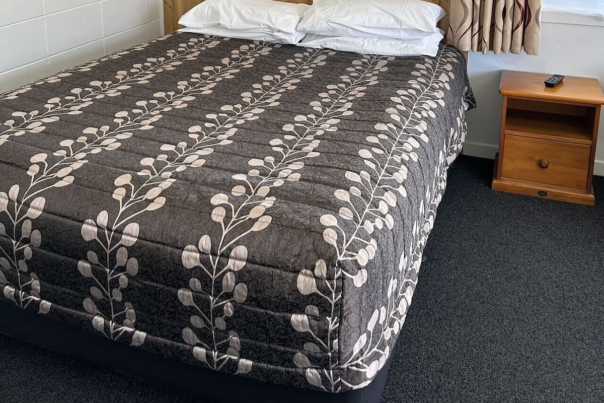 A hotel room with a bed, a wooden headboard, a nightstand with a remote control, and a window with curtains.