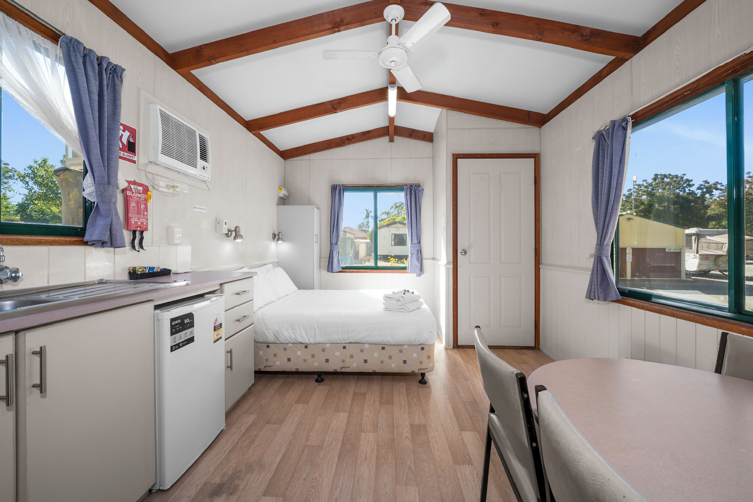 A compact room with a bed, a small kitchenette, a ceiling fan, and a window with curtains.