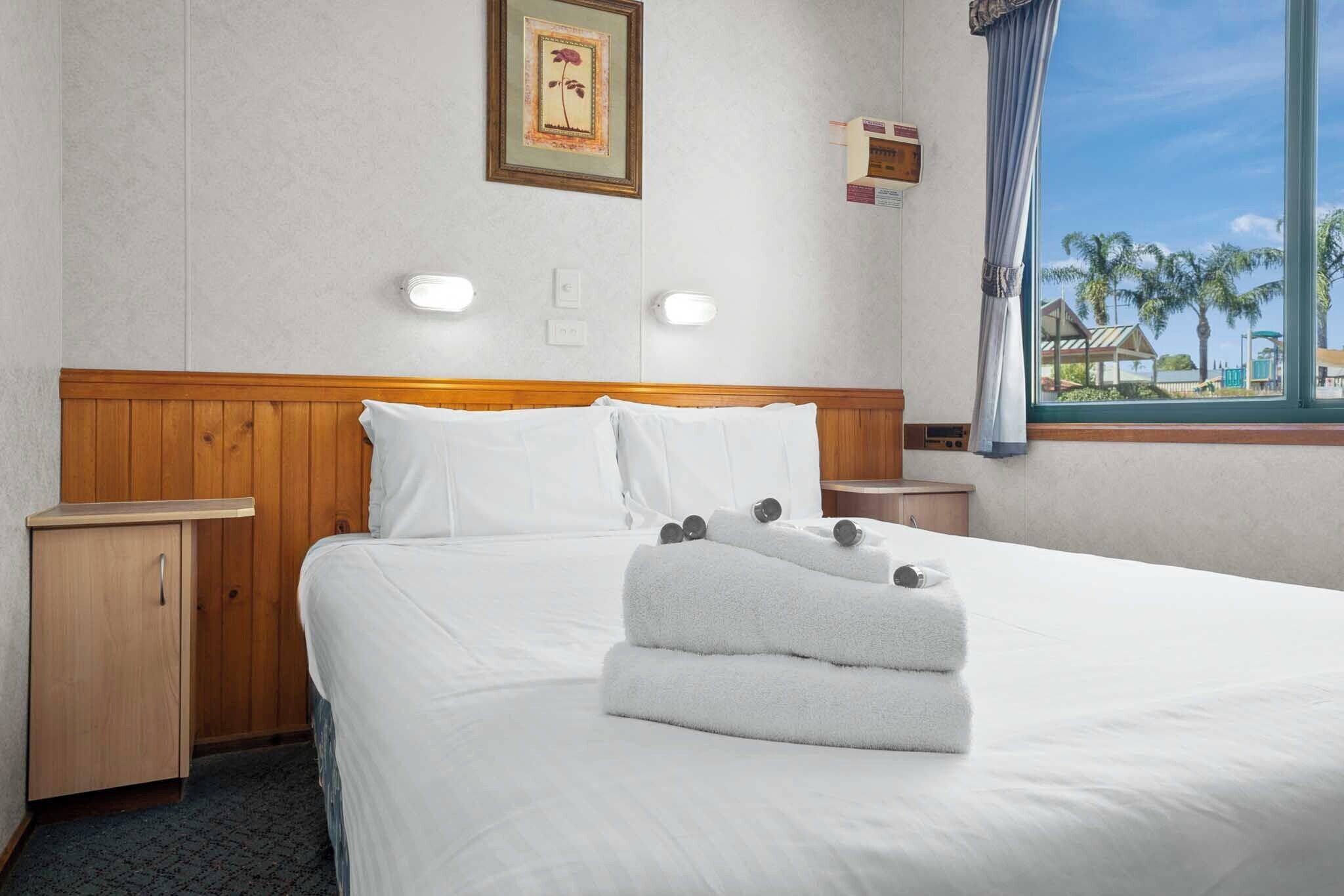 Habitación de hotel con cabecera de madera, ropa de cama blanca, cama bien hecha con toallas, mesita de noche, un mueble pequeño, un cuadro en la pared y ventana con vistas a palmeras y edificios.