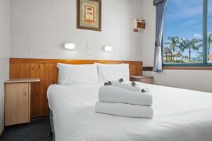 Habitación de hotel con cabecera de madera, ropa de cama blanca, cama bien hecha con toallas, mesita de noche, un mueble pequeño, un cuadro en la pared y ventana con vistas a palmeras y edificios.