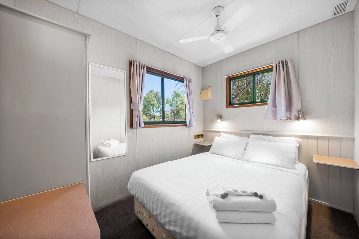 Une chambre d’hôtel avec un lit, une table de chevet, une chaise, une fenêtre avec des rideaux et un ventilateur de plafond.