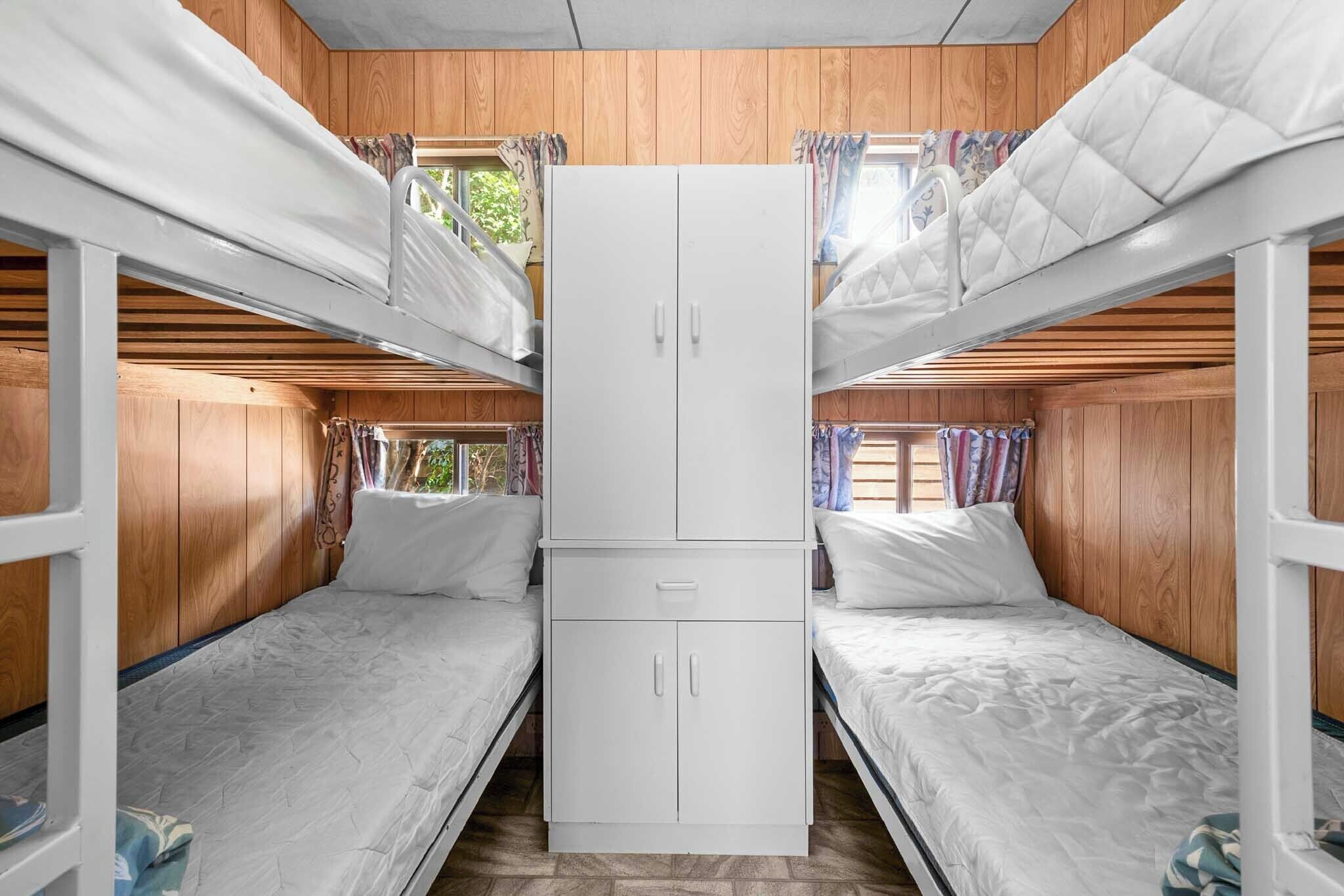 A bunk bed room with wooden walls, a central wardrobe, and a window with curtains.