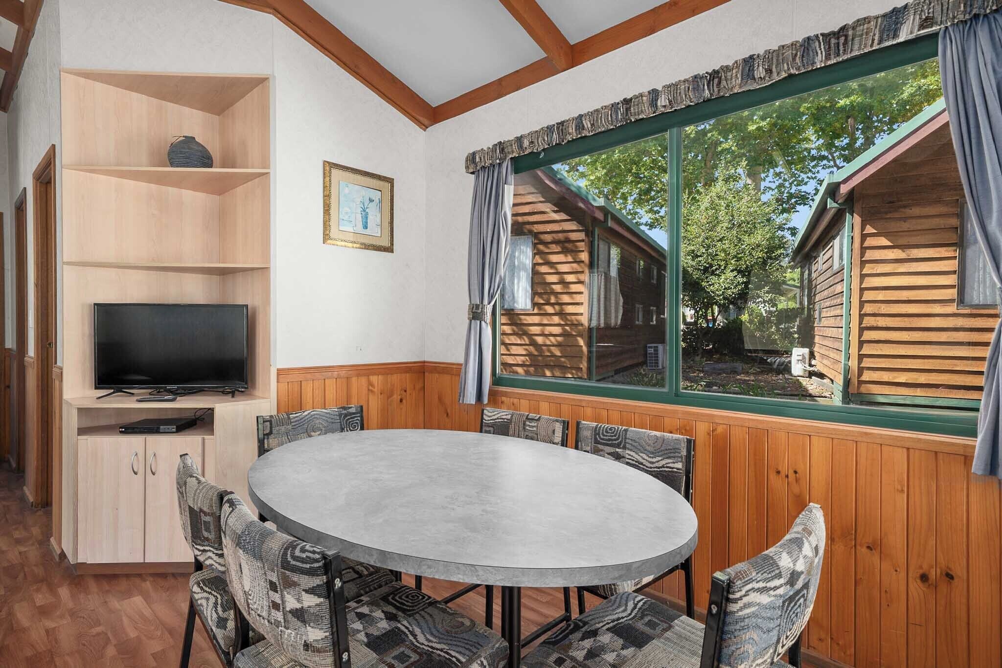 A cozy room with a round table, chairs, a TV, wooden shelves, and a large window with curtains.