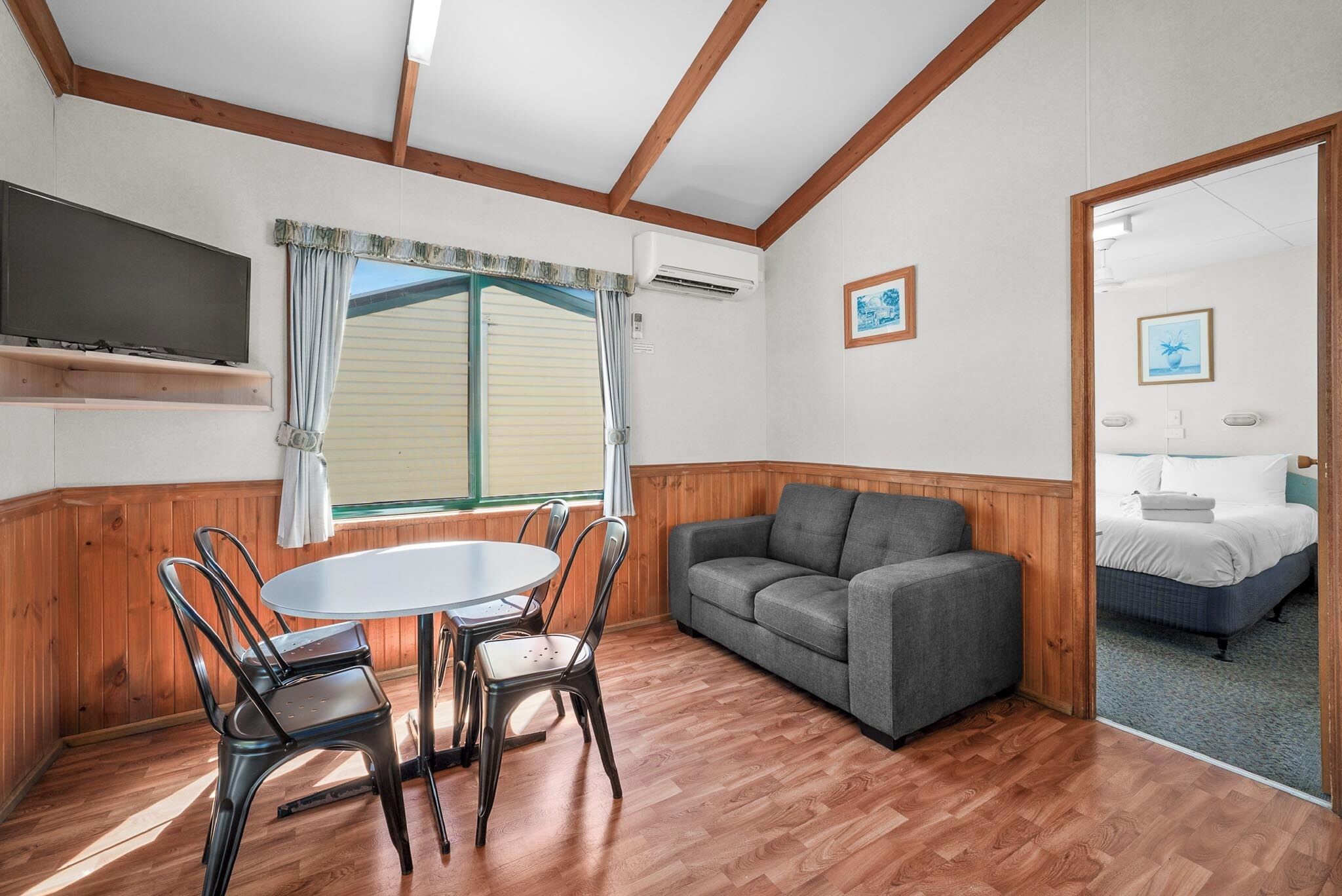 Une pièce avec un plafond en bois, une table ronde entourée de chaises, un canapé gris, un lit avec une literie blanche et un téléviseur fixé au mur.