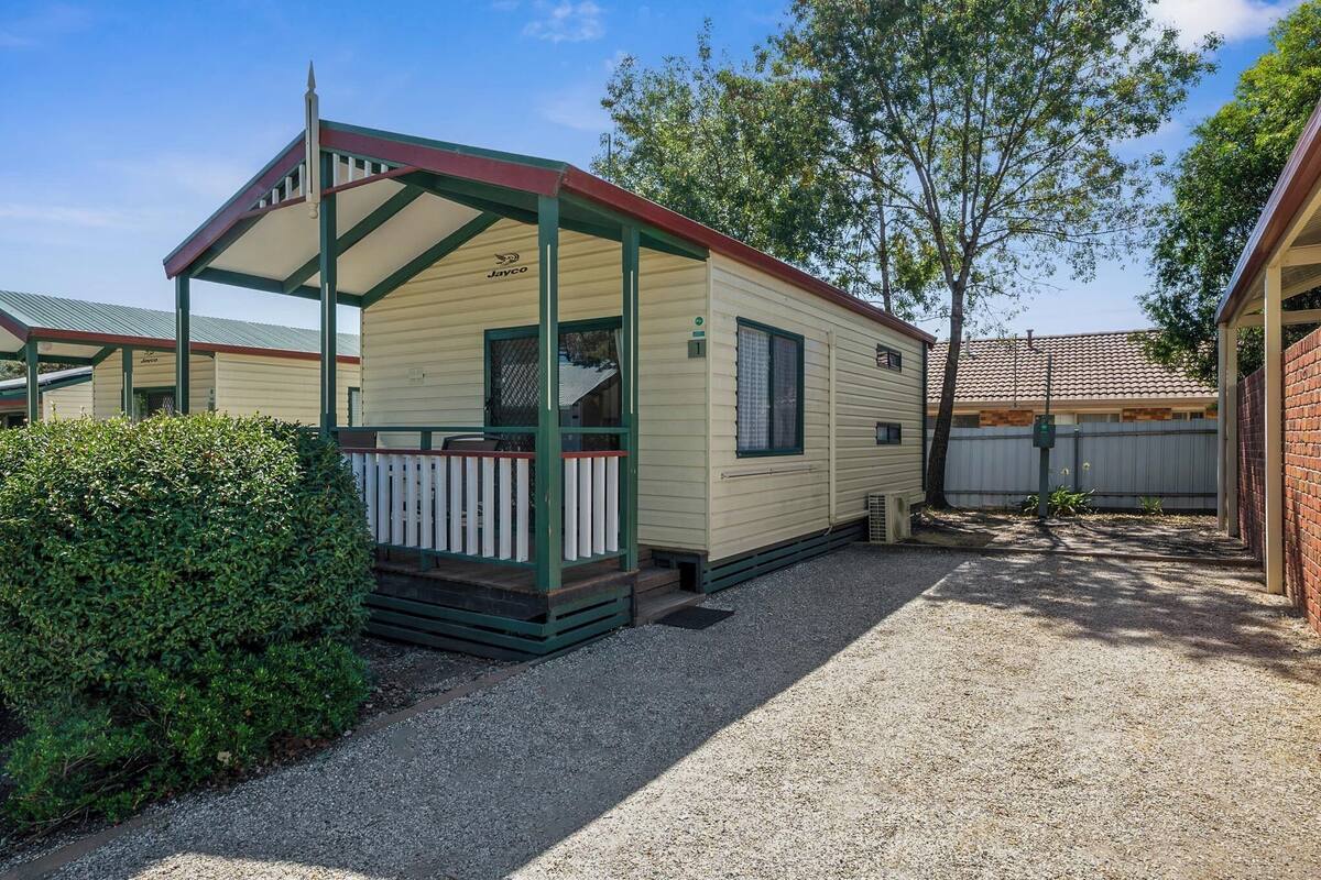 Une maison mobile dotée d’un porche couvert et d’une petite terrasse, entourée de gravier et de verdure.