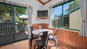 Una habitación con terraza de madera, una mesa redonda con cuatro sillas, un televisor montado en la pared y ventanas grandes con cortinas.