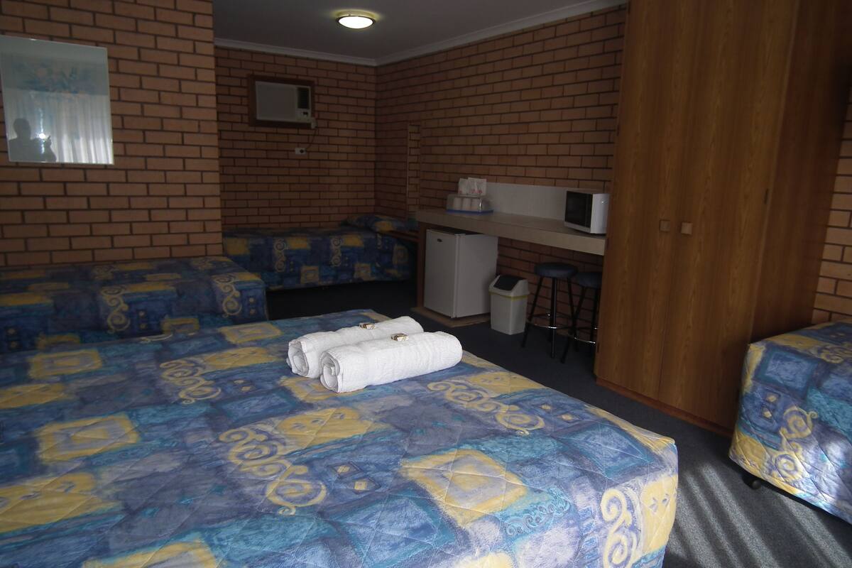 A hotel room with a brick wall, a bed with a patterned bedspread, a desk with a computer, and a small kitchenette.